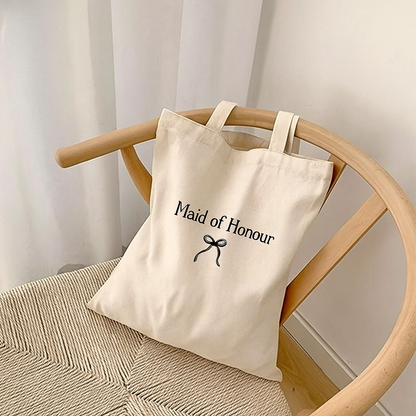 Beige tote bag with 'Maid of Honour' text on a wooden chair against a white curtain background