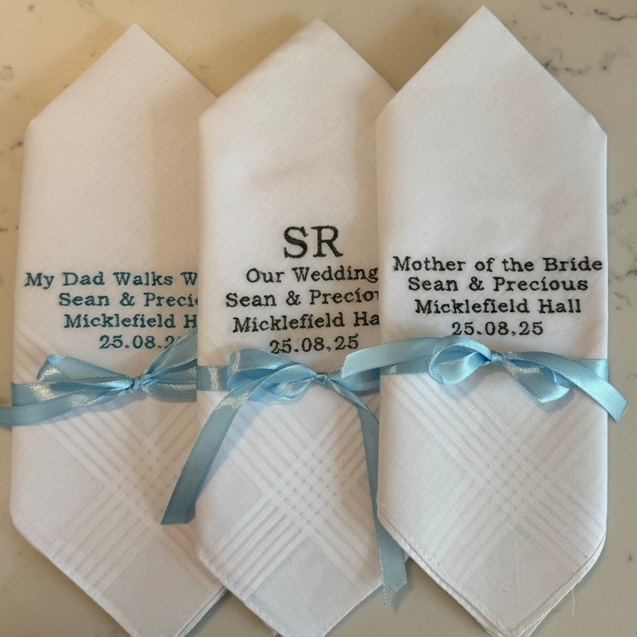 Three white handkerchiefs with personalized text and ribbons on a marble surface