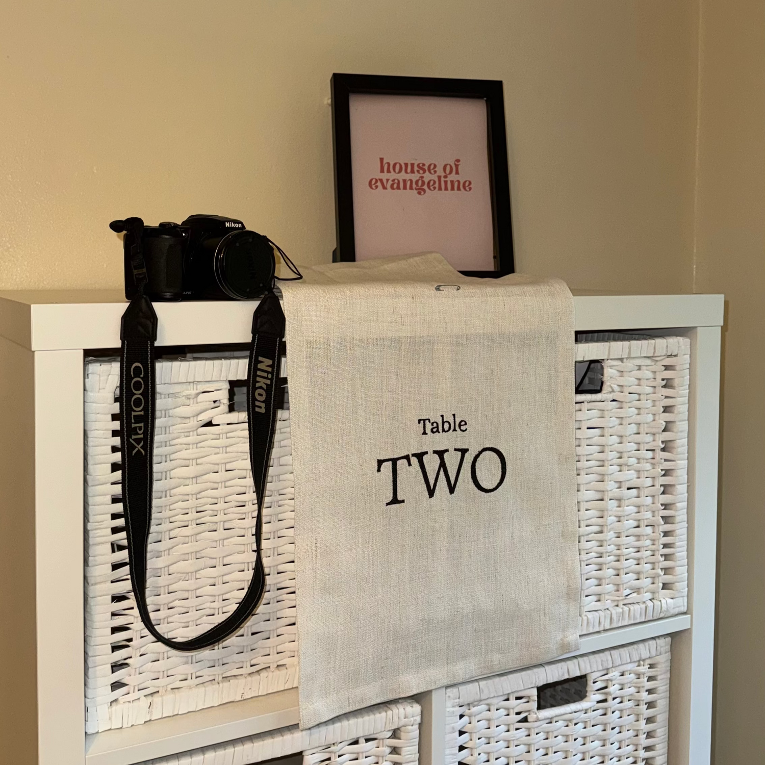 White wicker cabinet with a black camera and a tablecloth labeled 'Table Two' on top, against a beige wall.
