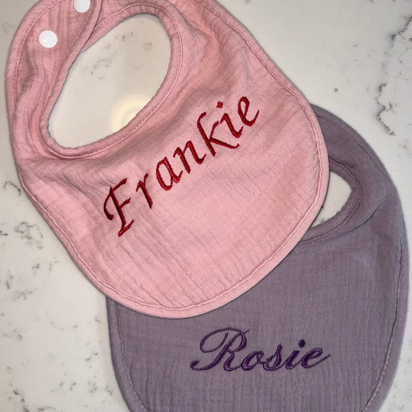 Pink bib with 'Frankie' embroidered and purple bib with 'Rosie' embroidered on a marble surface.