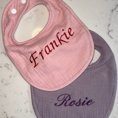 Pink bib with 'Frankie' embroidered and purple bib with 'Rosie' embroidered on a marble surface.