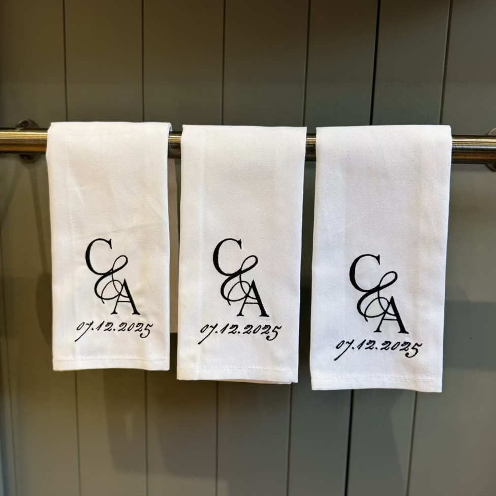 Three white towels with personalized monogram and date on a towel rack.