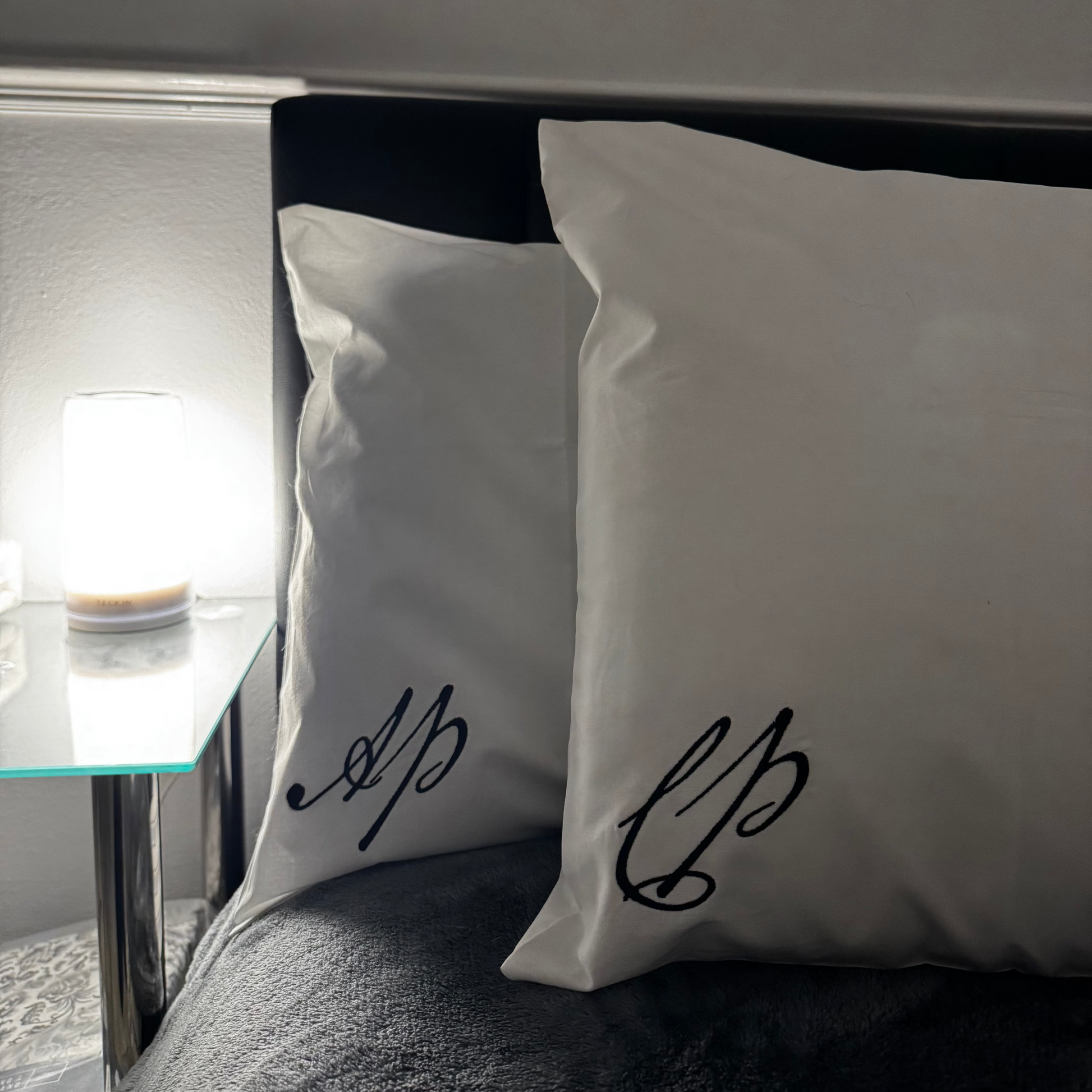 Two white pillows with black script on a bed next to a nightstand with a lamp.