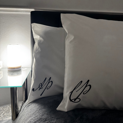 Two white pillows with black script on a bed next to a nightstand with a lamp.
