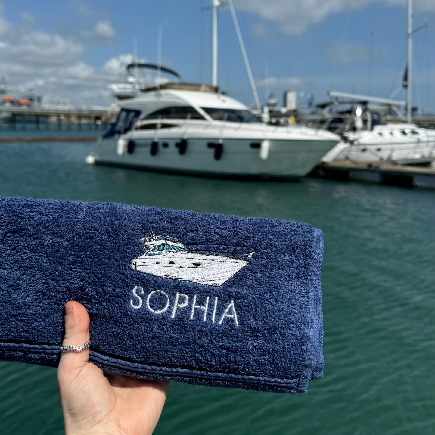 Navy blue towel with 'SOPHIA' and a logo held in front of a marina with boats.